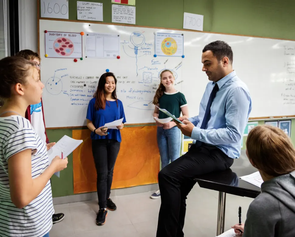 Teacher sitting on desk with Kids standing around the room listening